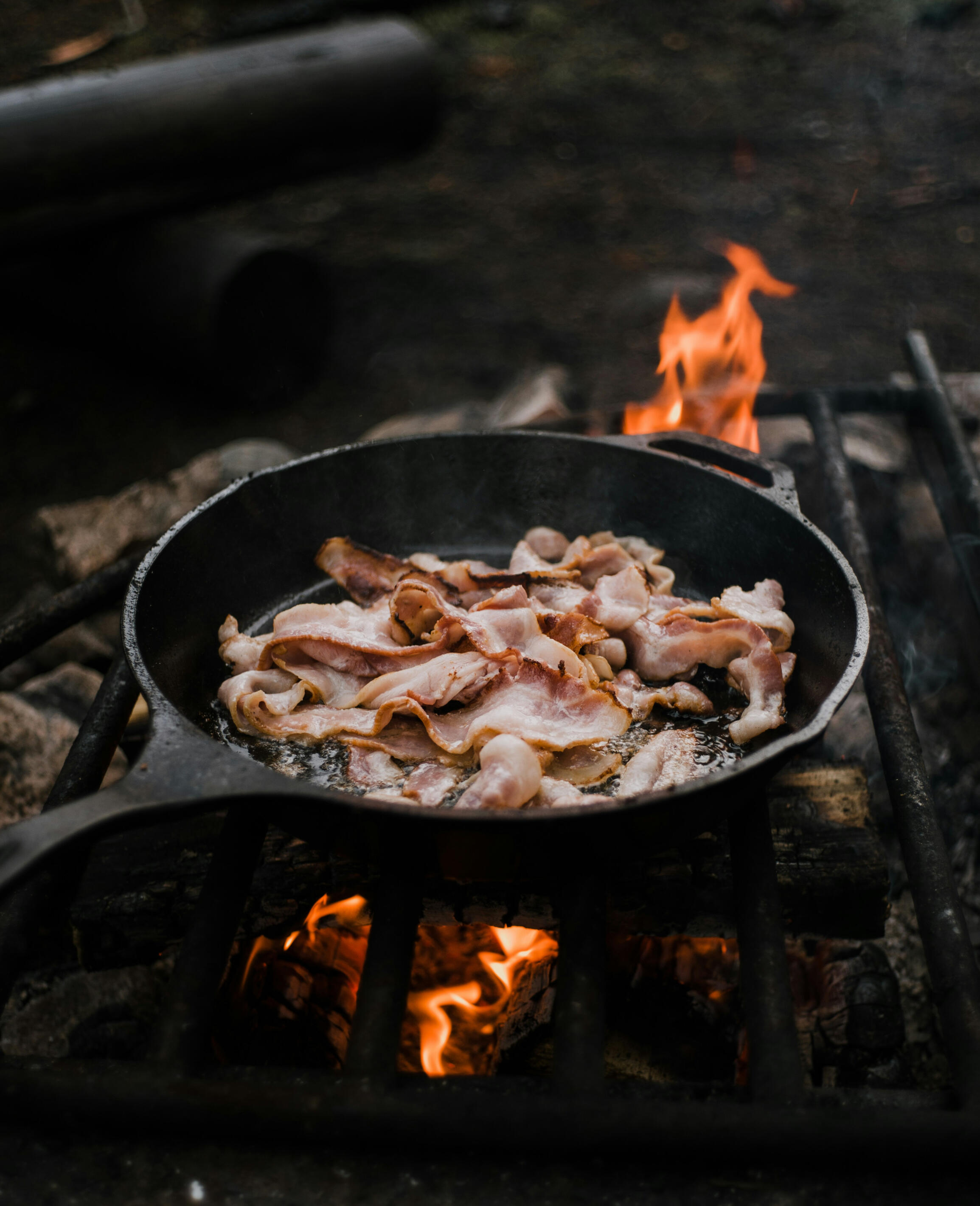 Cooking over open flame. Swedish Wild Kitchen, Åsensbruk Strips of bacon sizzling in a cast iron pan over a wood fire. Swedish Wild Kitchen, Åsensbruk.