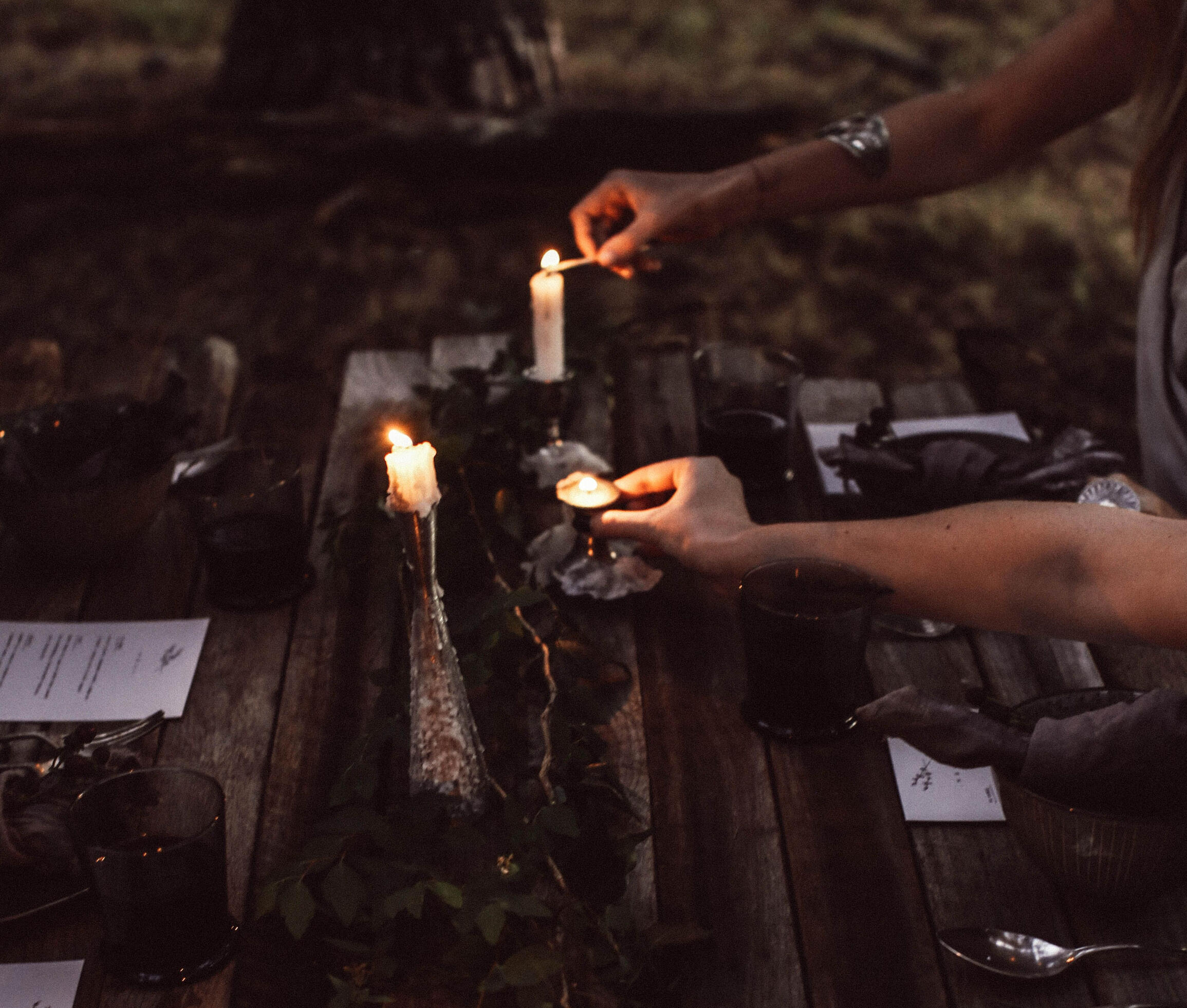 Outdoor dining by candlelight. Swedish Wild KItchen, Åsensbruk Rustic wooden table set outdoors with candles, glasses, and a hand lighting a small candle.
