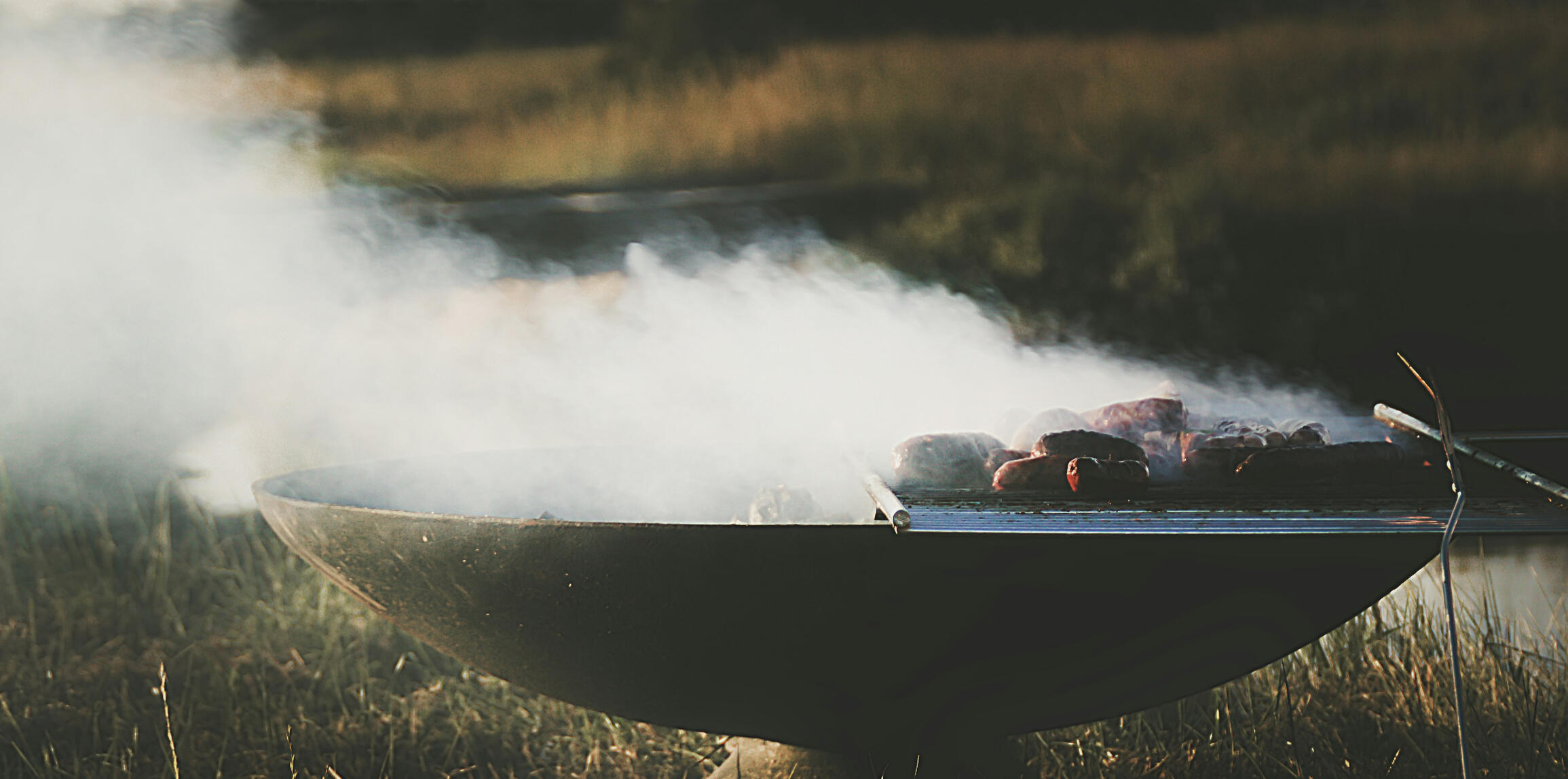 Smoke rising from the grill. Swedish Wild Kitchen, Åsensbruk Meat cooking on a round outdoor grill with thick smoke rising over grass. Swedish Wild Kitchen, Åsensbruk